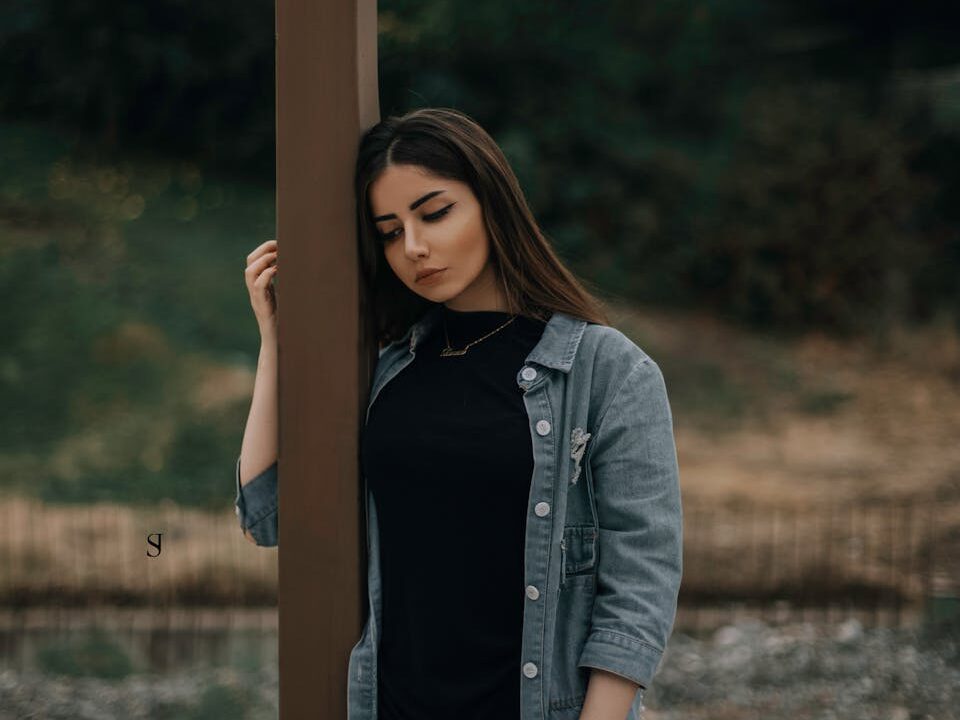 Young woman in a denim jacket standing outdoors, deep in thought.