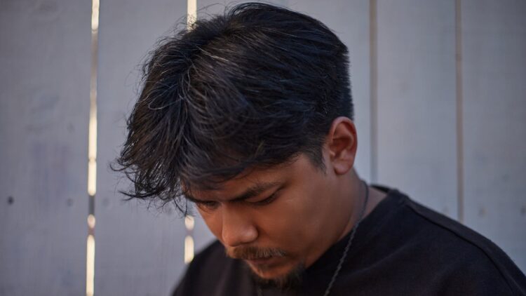Close-up of a young man with thoughtful expression against a wooden fence background.