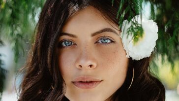 Close-up portrait of a woman with blue eyes and dark hair wearing a white flower outdoors.