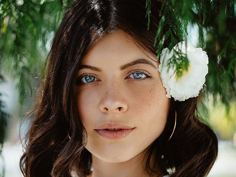 Close-up portrait of a woman with blue eyes and dark hair wearing a white flower outdoors.