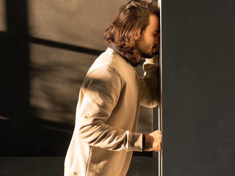 A man in casual attire showing frustration by leaning against a wall in a dark room.