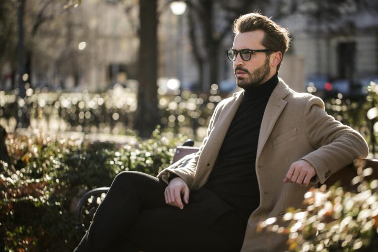 A fashionable man with a beard and eyeglasses sits on a park bench during the day.