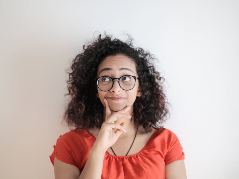 Portrait of a thoughtful woman in a red top, with curly hair and glasses against a white background.