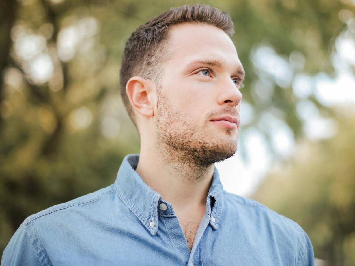 Portrait of a handsome young man with a beard looking thoughtfully in a sunny park setting.
