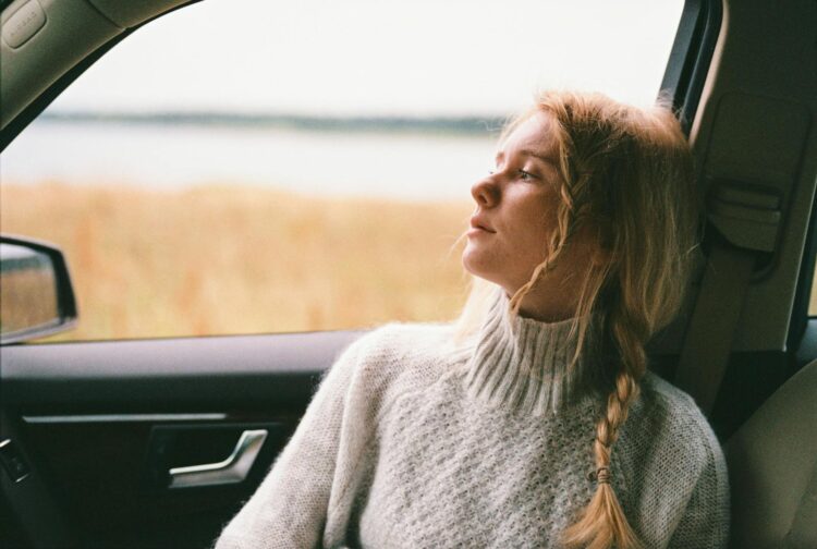A thoughtful woman with braided hair gazes out a car window during a daytime road trip.