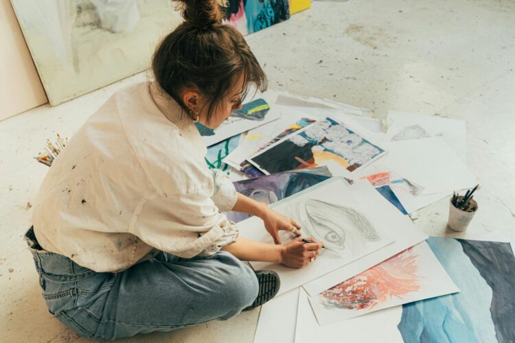 A young woman sketching on the floor surrounded by her artistic drawings in a creative studio.