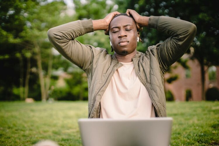 An upset man with closed eyes holds his head while using a laptop outdoors.