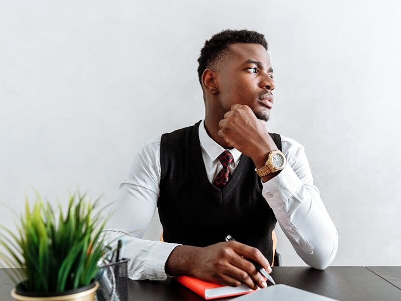 Businessman in formal attire sitting at a conference table with laptop and plant, looking thoughtful.
