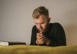 Bearded man in a knitted sweater praying with a Bible beside him in a serene indoor setting.