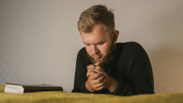 Bearded man in a knitted sweater praying with a Bible beside him in a serene indoor setting.