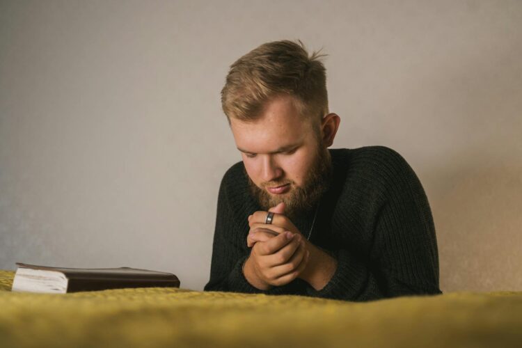 Bearded man in a knitted sweater praying with a Bible beside him in a serene indoor setting.