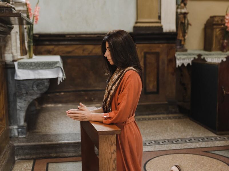 A woman kneels in prayer at a wooden kneeler inside a church.