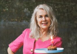 A joyful senior woman in a pink dress carrying a lit birthday cake in an indoor setting.