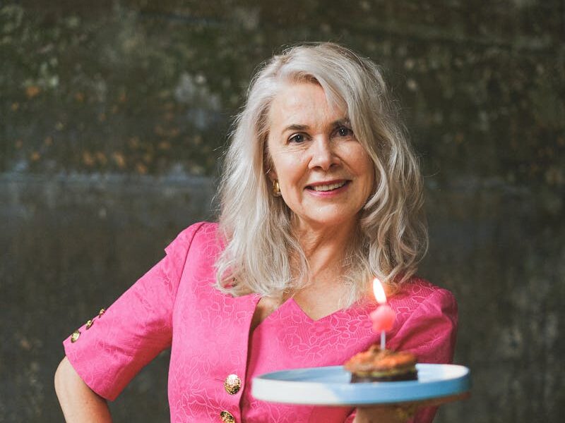 A joyful senior woman in a pink dress carrying a lit birthday cake in an indoor setting.