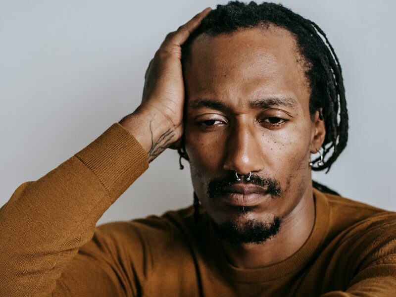 A man in a brown sweater sits pensively, resting his head on his hand, conveying emotion.