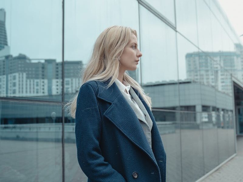 Confident woman in a blue coat stands by a reflective glass building, gazing thoughtfully.