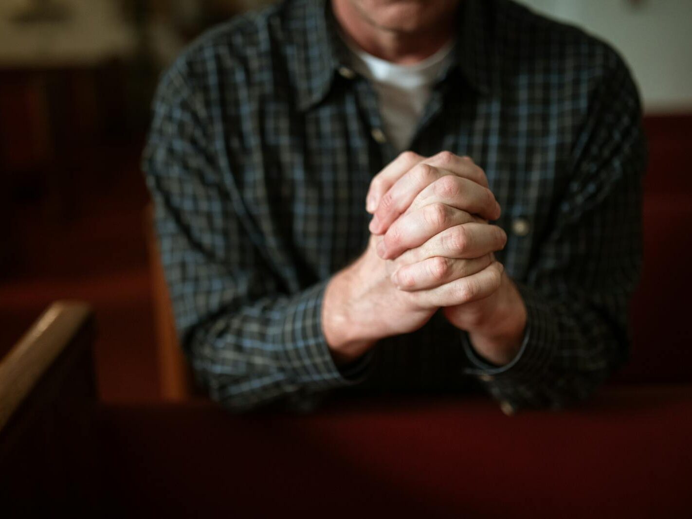A man in a checked shirt praying in a church, hands clasped together.