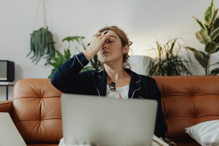 Stressed woman sitting with a drink, laptop, and plants around, expressing exhaustion.