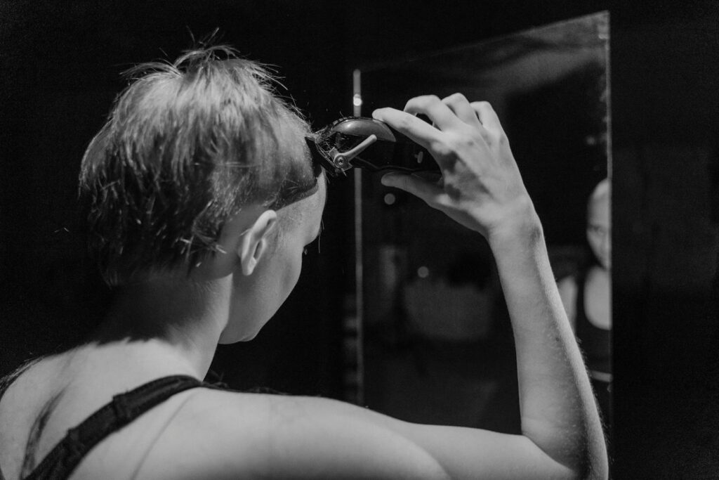 Black and white image of a woman using clippers to shave her head, reflected in a mirror.