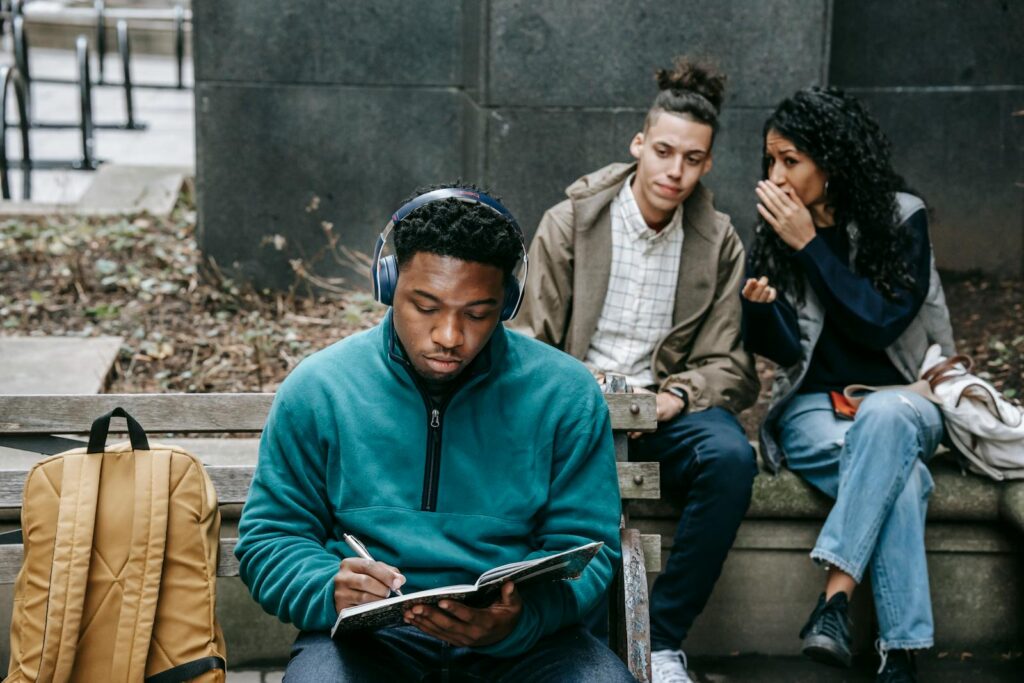 Young multiethnic friends in casual outfit with backpack gossiping behind back of African American guy sitting on bench in headphones and taking notes in notebook in city street in daylight