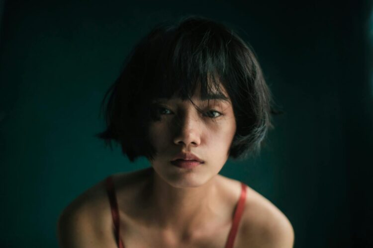 Sad young Asian female in red top looking at camera in studio with green wall