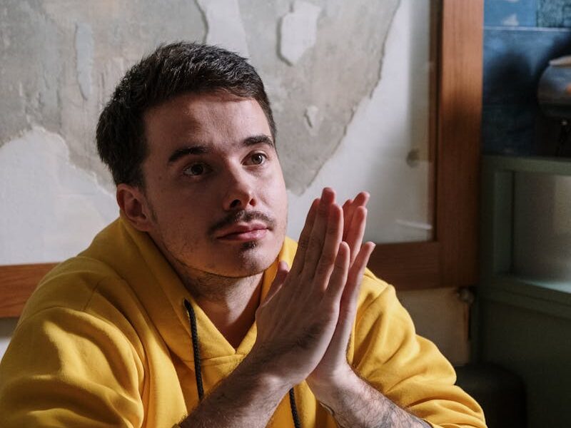 Man in yellow hoodie thinking deeply next to a laptop indoors.