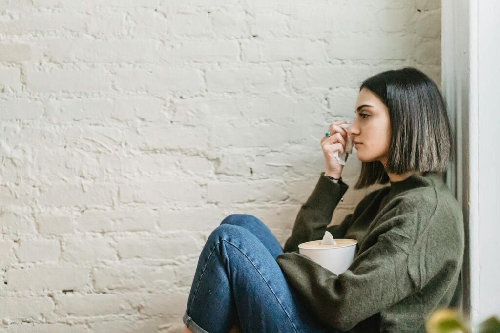 Side view of depressed young ethnic female millennial in warm sweater sitting leaning on wall and wiping tears with tissue at home