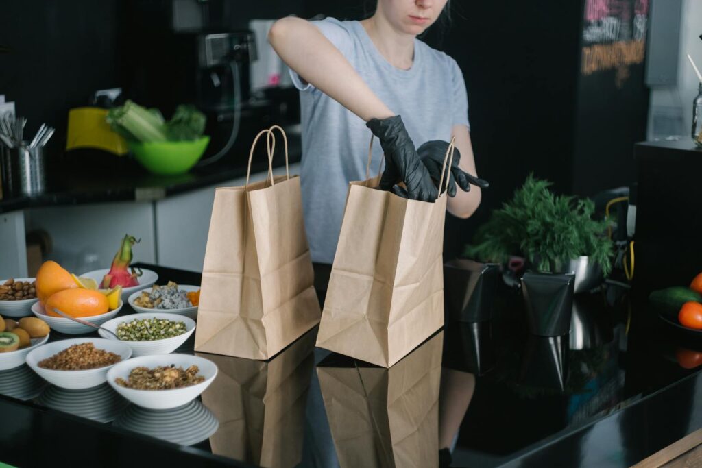 A person in a kitchen packing food ingredients into paper bags for takeout orders.