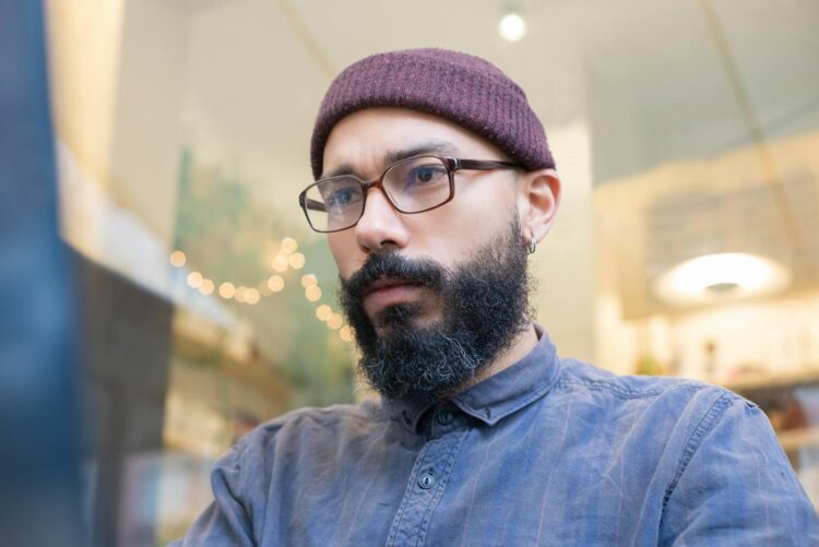 Focused portrait of a bearded man with glasses and beanie in a cozy cafe setting.