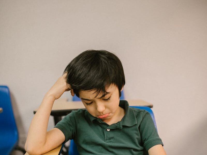 A young boy sits alone at a desk, looking sad and thoughtful in a classroom setting.