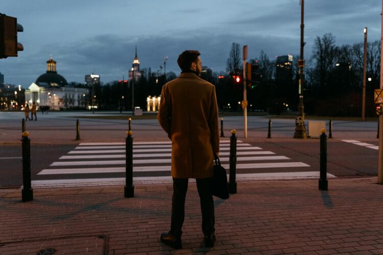 An adult man in a coat waits at a pedestrian crossing during a city evening.