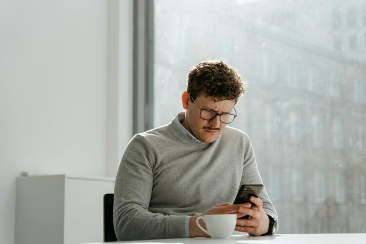 Adult man wearing glasses using smartphone in sunny office space, holding a mug.