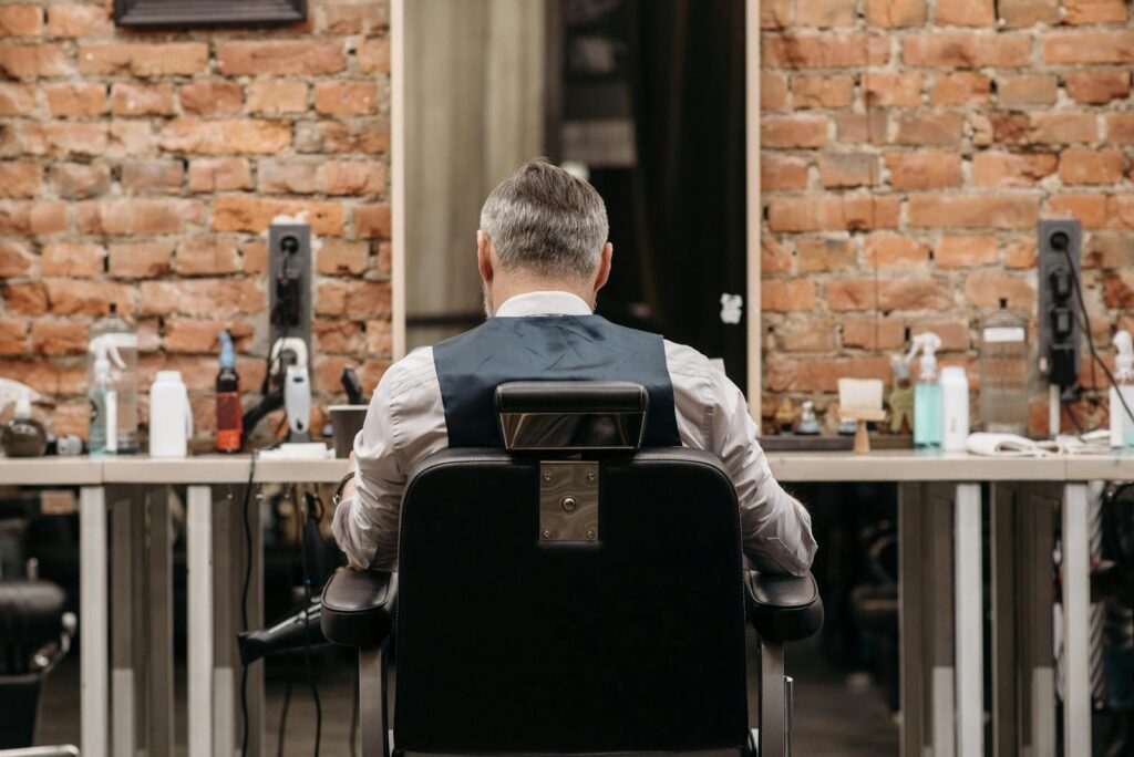 Backview of a man in a barbershop, sitting on a leather seat, facing a mirror against a brick wall.
