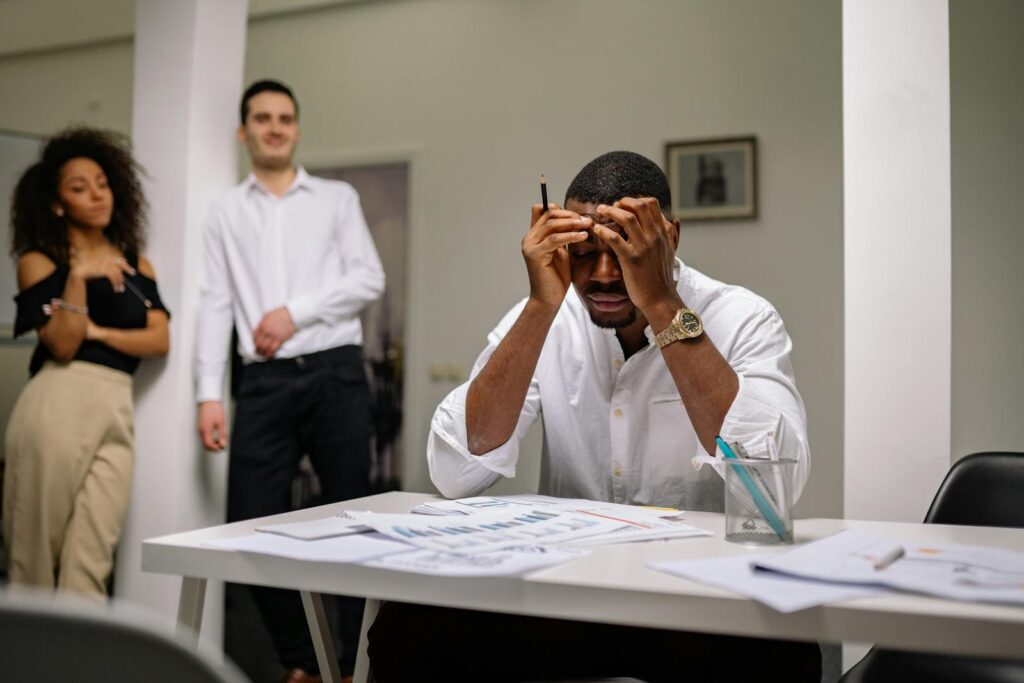 Stressed businessman sitting with hands on head while colleagues stand nearby in office.