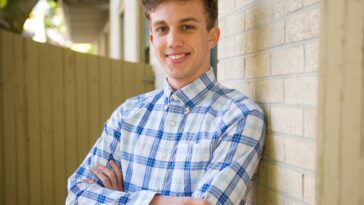 Casually dressed young man smiling, leaning against a brick wall outdoors on a sunny day.