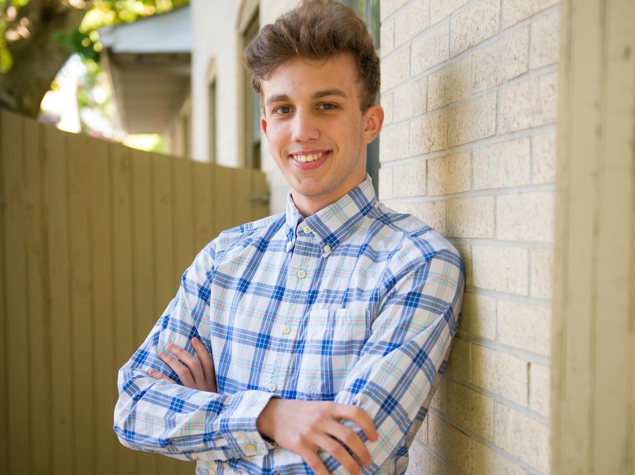 Casually dressed young man smiling, leaning against a brick wall outdoors on a sunny day.