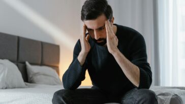 A man in a black sweater sits on a bed, expressing frustration in a bedroom.