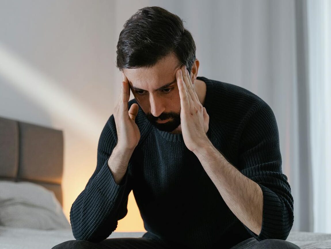A man in a black sweater sits on a bed, expressing frustration in a bedroom.