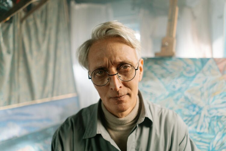 Portrait of a senior artist in glasses at his studio, surrounded by abstract art.