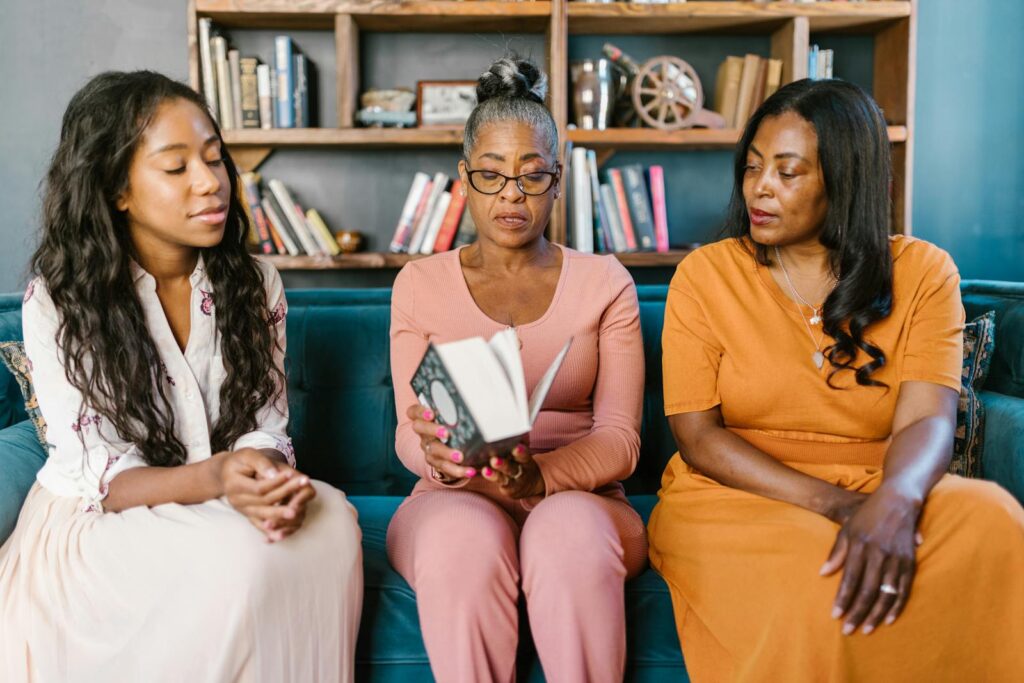 Three women sitting on a couch reading a book together in a cozy library setting.