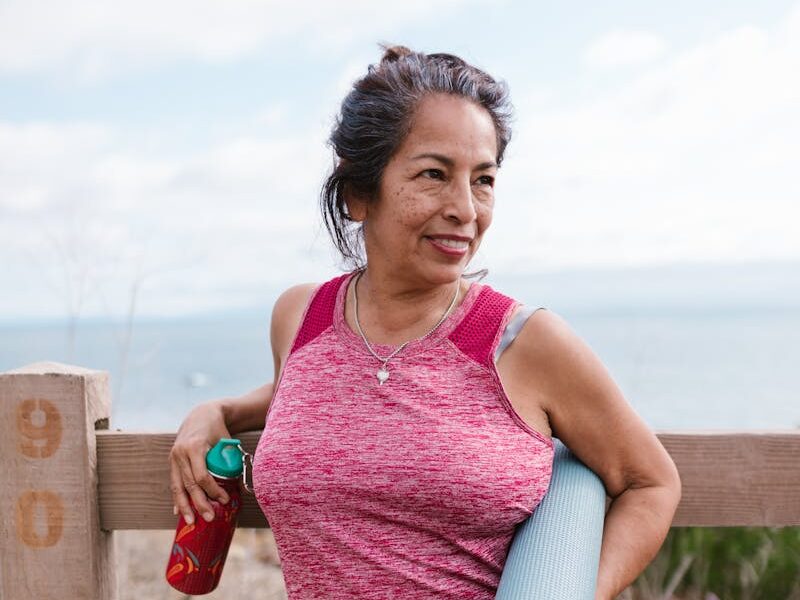 Smiling senior woman holding yoga mat outdoors by the beach, embracing active lifestyle.