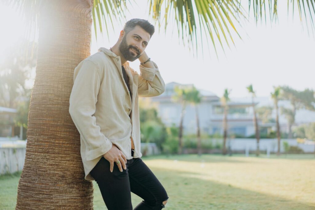 Stylish man smiling while leaning on a palm tree in a sunny outdoor garden setting.