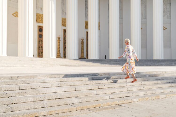 Senior woman in floral dress walking up outdoor stairs beside grand columns.