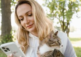 Smiling woman with cat using phone outdoors on a sunny day.