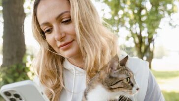 Smiling woman with cat using phone outdoors on a sunny day.