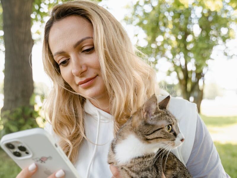 Smiling woman with cat using phone outdoors on a sunny day.