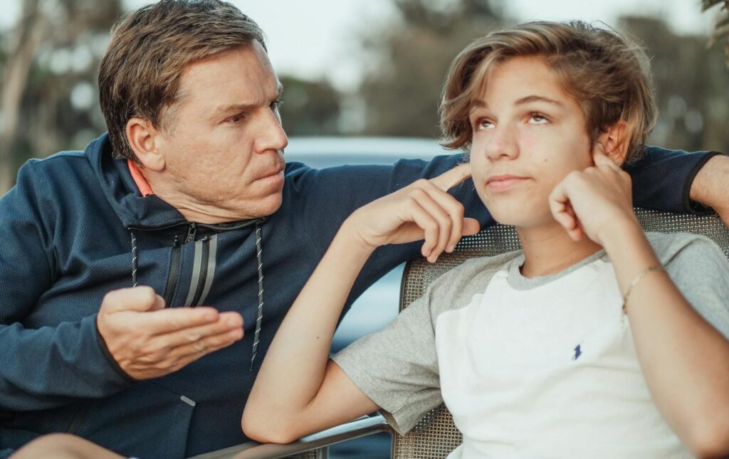 Father expressing frustration while teenage son shows defiance, seated outdoors.