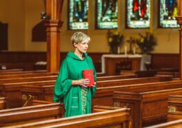 A female priest in green chasuble holds a Bible while walking through a church aisle.