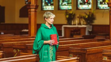 A female priest in green chasuble holds a Bible while walking through a church aisle.