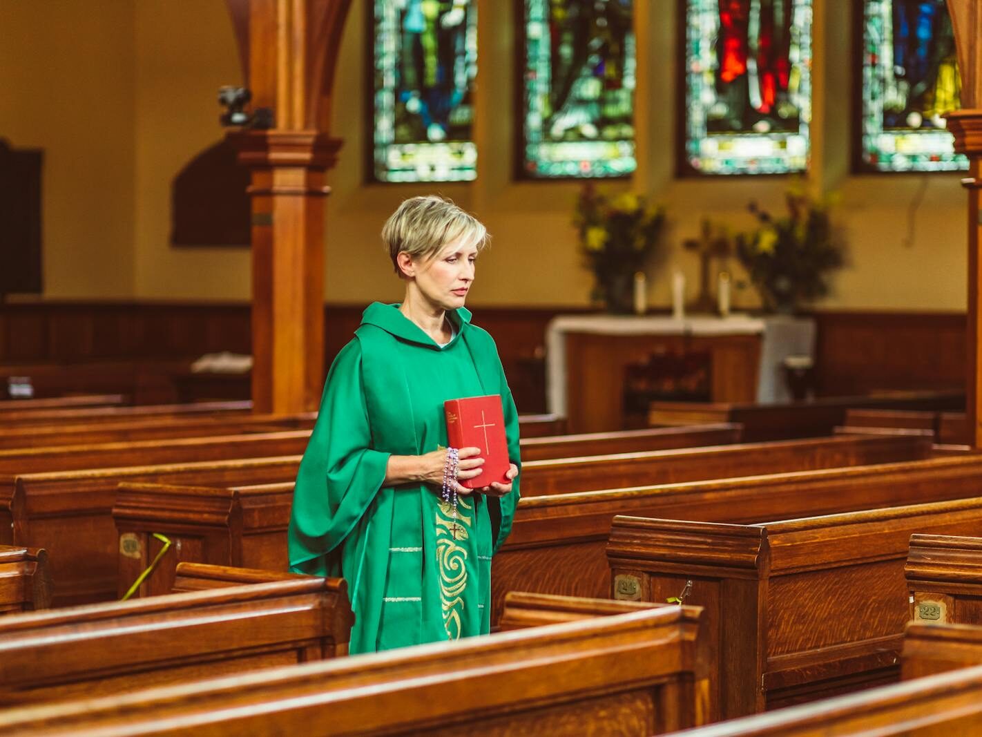 A female priest in green chasuble holds a Bible while walking through a church aisle.